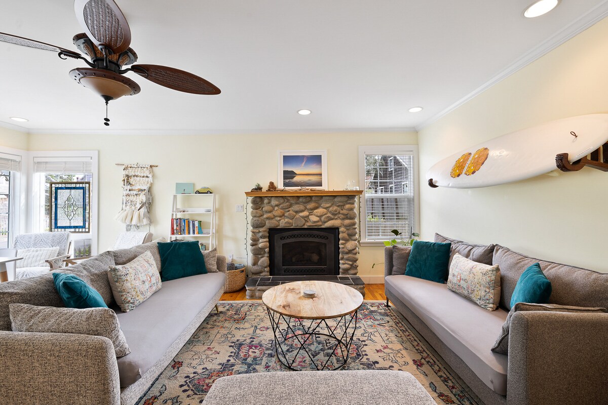 A spacious living room features a stone fireplace as the focal point, surrounded by two large sofas with decorative pillows. A wooden coffee table is centered on a colorful area rug, with natural light filtering through the nearby windows. A surfboard is mounted on the wall.