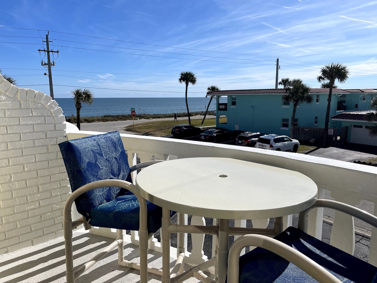 A balcony area is seen with a round table and two chairs featuring blue fabric. The ocean is visible in the background, framed by palm trees and the clear sky, creating a serene seaside atmosphere.