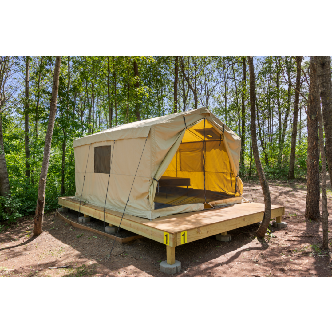 A sturdy cabin tent is set on a wooden platform surrounded by trees. The tent, made of light-colored canvas, features large front openings with screens, allowing for natural light. The structure is anchored with ropes and positioned on elevated supports for stability.