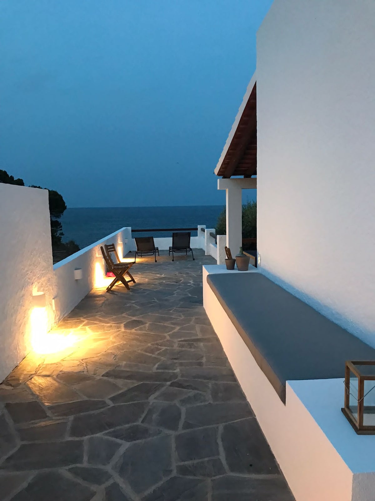 A tranquil outdoor terrace features a stone pathway leading to ocean views. Soft lighting illuminates the space, highlighting a seating area with lounge chairs. A white wall provides a clean contrast against the serene backdrop of the sea, creating an inviting atmosphere for relaxation.