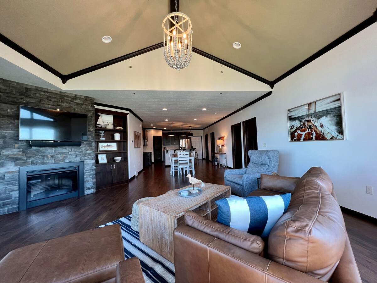 The living area features vaulted ceilings and a stone accent wall, with a large television mounted above a contemporary fireplace. Comfortable seating is presented in earthy tones, complemented by a light wood coffee table. A dining area is visible in the background, promoting an open layout.
