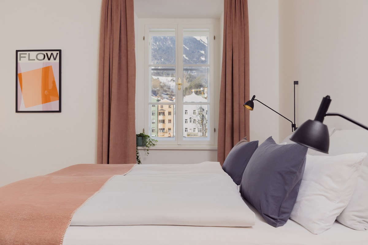 A cozy bedroom features a spacious bed adorned with a soft blanket and two decorative pillows. A window allows natural light to enter, offering a view of the mountains. A contemporary art piece hangs on the wall, and a bedside lamp adds functionality.