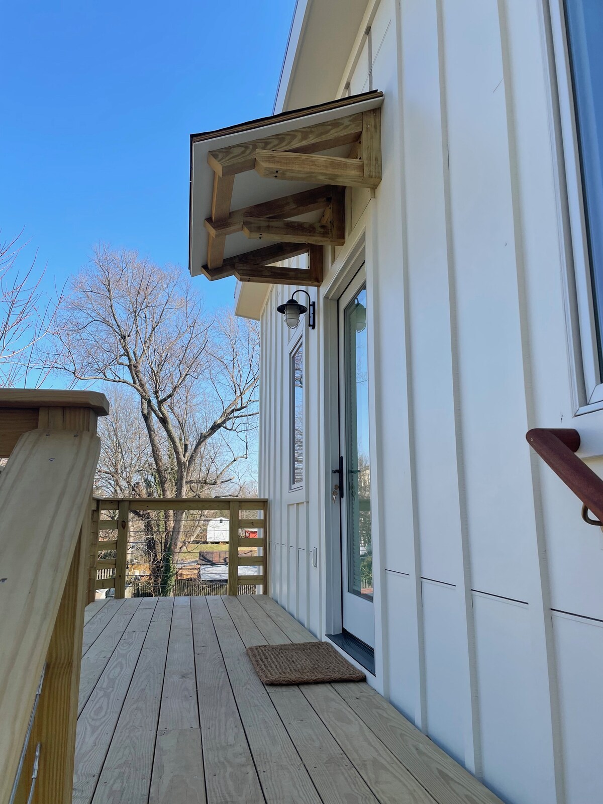 The exterior entrance is framed by a wooden overhang, providing shelter from the elements. A cozy mat lays at the door, and the wooden deck connects to a railing, enhancing the welcoming feel of the space. Surrounding trees are visible against the clear blue sky.