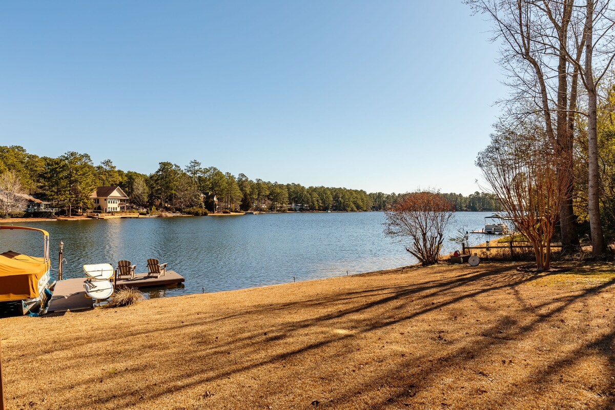 A serene lakeside view captures the calm waters of Lake Pinehurst, bordered by gently sloping grass. Bare trees cast shadows along the shore, while neighboring houses are visible in the distance. A small dock and boats are stationed at the water's edge, inviting relaxation.