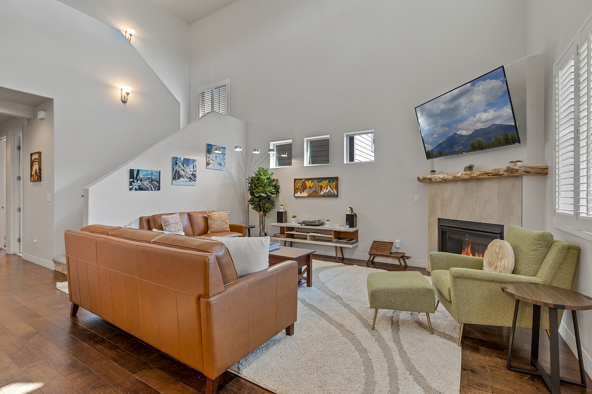 An open-plan living area features two comfortable sofas and modern armchairs arranged around a stylish coffee table. A smart TV is mounted above a cozy indoor fireplace, and large windows allow natural light to fill the space. Artwork decorates the walls, enhancing the welcoming atmosphere.