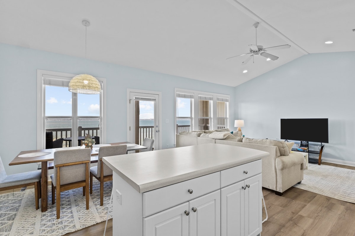 An open-concept living space is displayed, featuring a bright dining area with a wooden table surrounded by six chairs. A cozy seating arrangement is visible in the background, complemented by large windows offering views of the surrounding landscape.