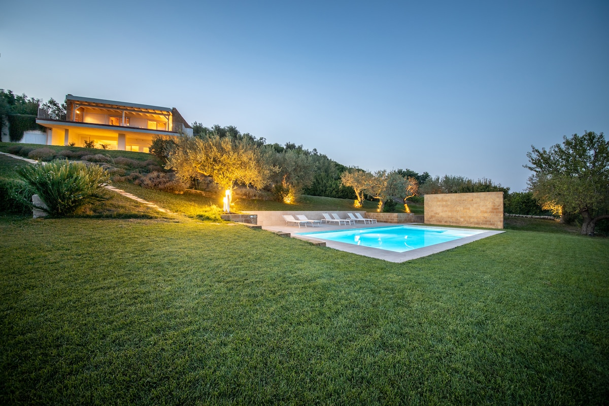 An outdoor view reveals a well-maintained lawn surrounding a swimming pool, illuminated by soft lighting. Olive trees and shrubbery frame the area, with a modern house visible in the background, showcasing large windows that reflect sunlight from the serene evening sky.