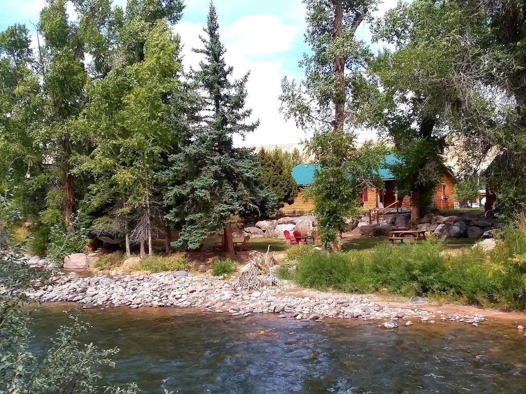 A vintage log cabin is nestled among tall trees along the Wind River, with a green roof complementing the natural surroundings. A rocky shoreline leads to a gently flowing river, while a small picnic area is visible, featuring a table and chairs beside the water.