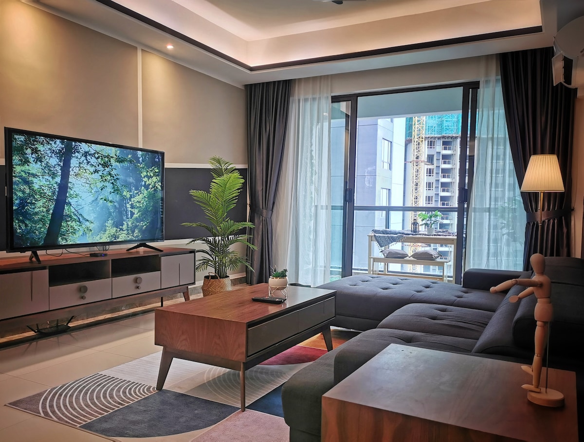 A comfortable living area features a large smart TV positioned on a modern entertainment unit. A plush sectional sofa invites relaxation, and a small dining table for two is visible on the balcony. Natural light fills the space through tall glass doors, creating a welcoming environment.