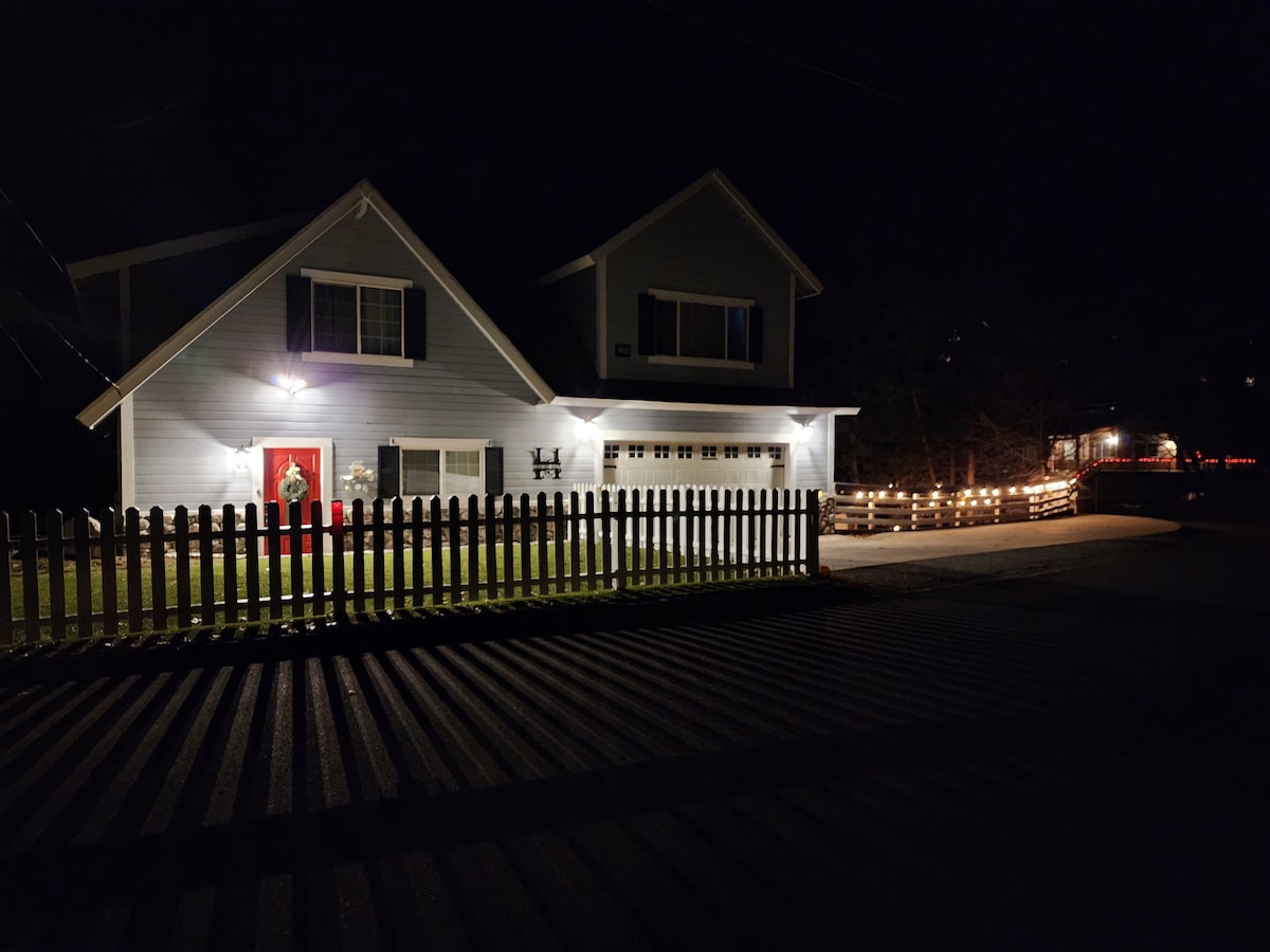 The exterior of a two-story house is depicted at night, featuring soft outdoor lighting that highlights the welcoming entry. A white picket fence surrounds the yard, with a well-lit driveway leading up to the garage. Nearby, the ambiance of ambient night lighting enhances the serene setting.