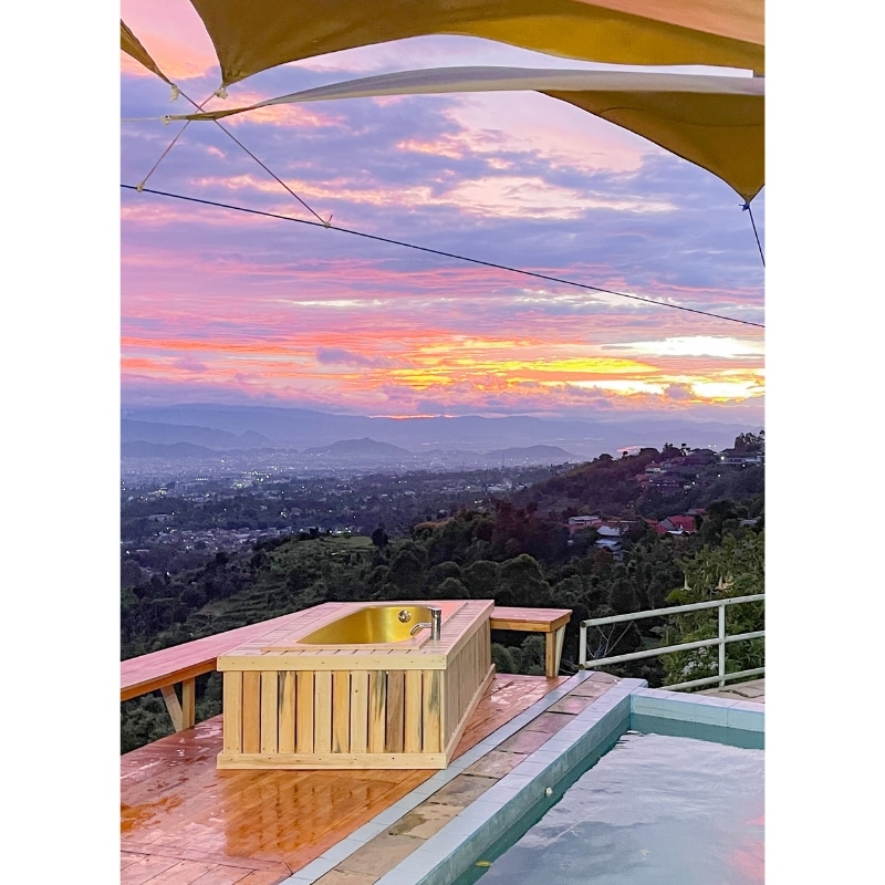A wooden hot tub overlooks a vibrant sunset, with mountains visible in the distance. The setting features a spacious deck and a pool nearby, creating an inviting space for relaxation and enjoying natural beauty.