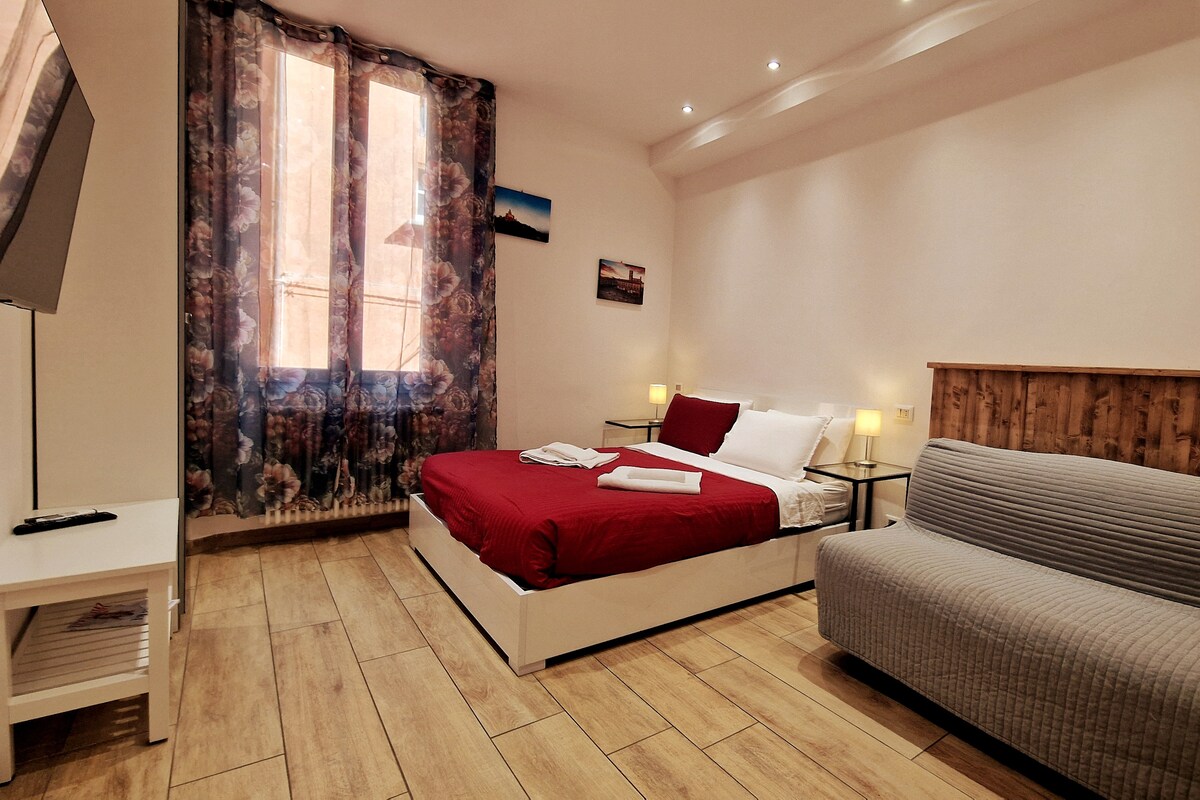 A bright sleeping area features a spacious bed dressed in red linens, complemented by white towels neatly arranged. Soft lighting is provided by bedside lamps, and a gray sofa is positioned against the wall. Two windows with floral curtains allow natural light to fill the room.