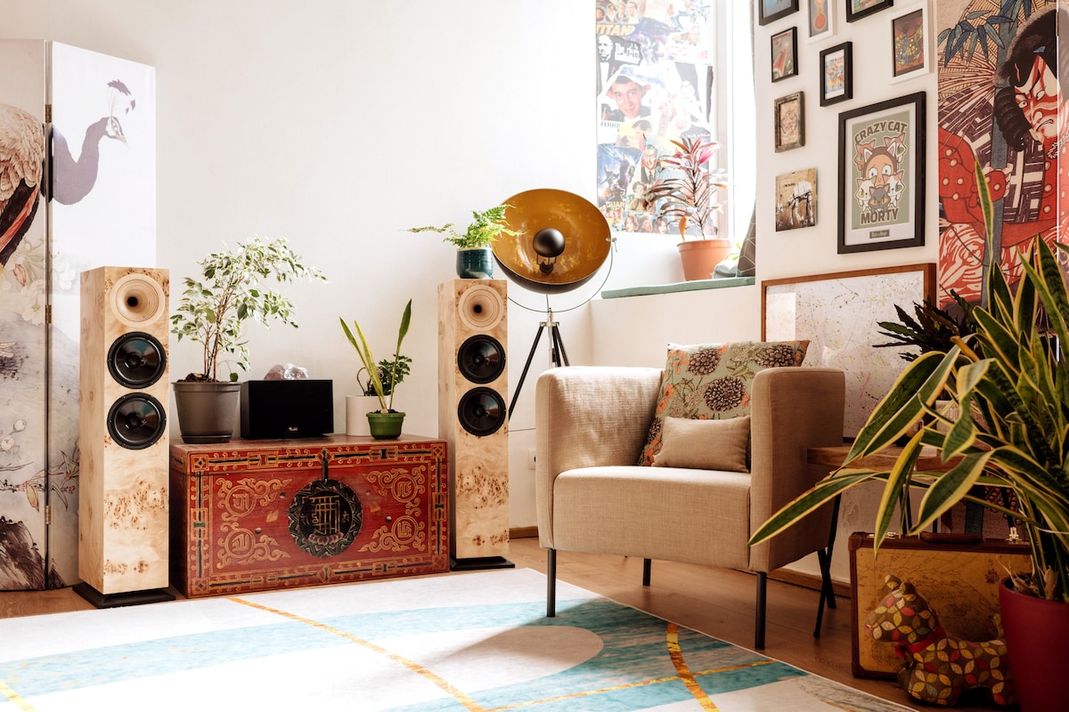 A modern living area features a comfortable chair alongside a decorative chest, with tall speakers positioned nearby. A vintage-style floor lamp illuminates the space, while green plants and an array of framed artwork add visual interest. A colorful area rug enhances the inviting atmosphere.
