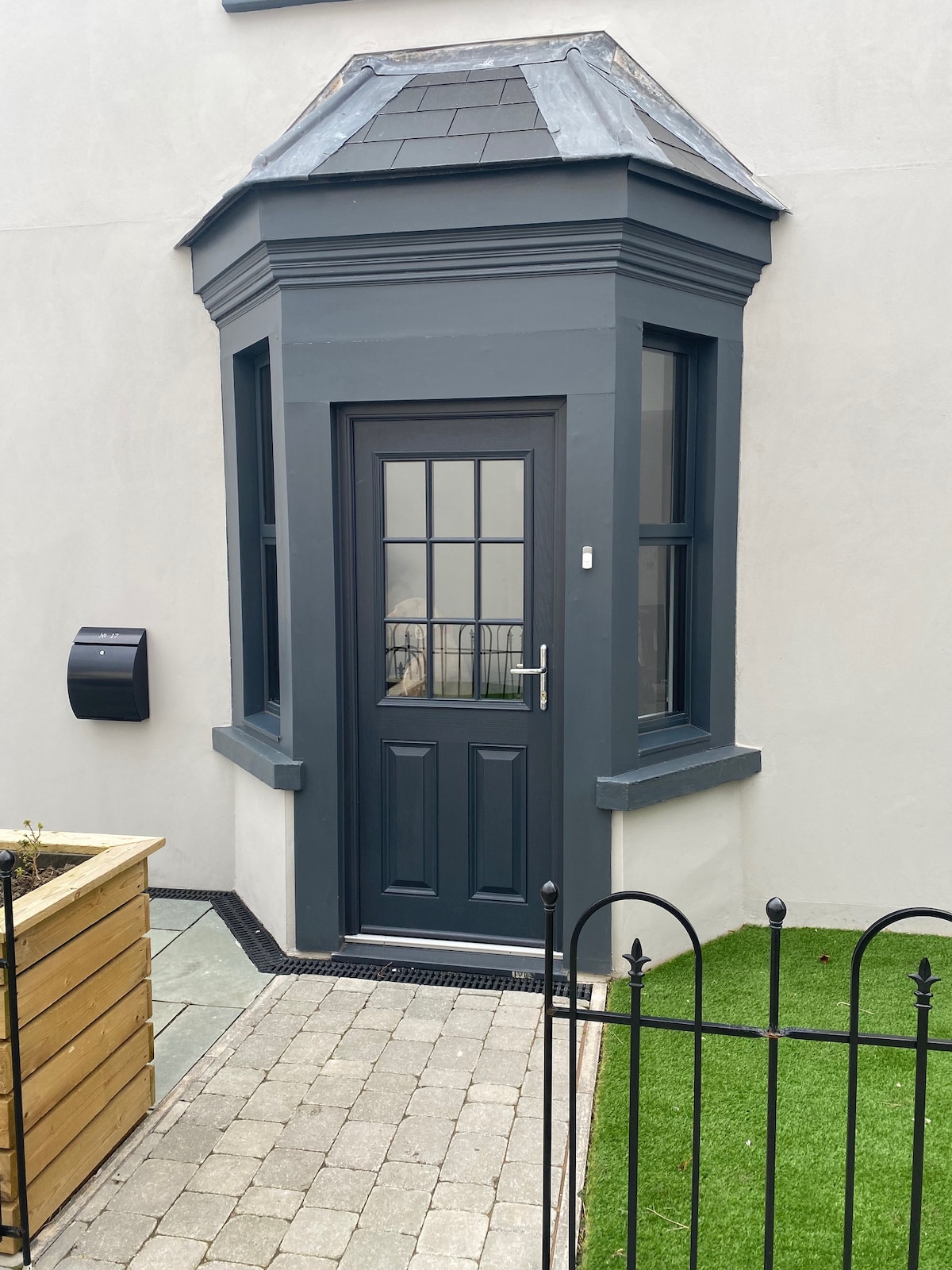 A stylish entrance is framed by a gray portico with a pitched roof. The door features a grid of glass panes, allowing natural light to enter. Surrounding the entryway are neatly arranged paving stones and a small patch of green grass, enhancing the outdoor space.