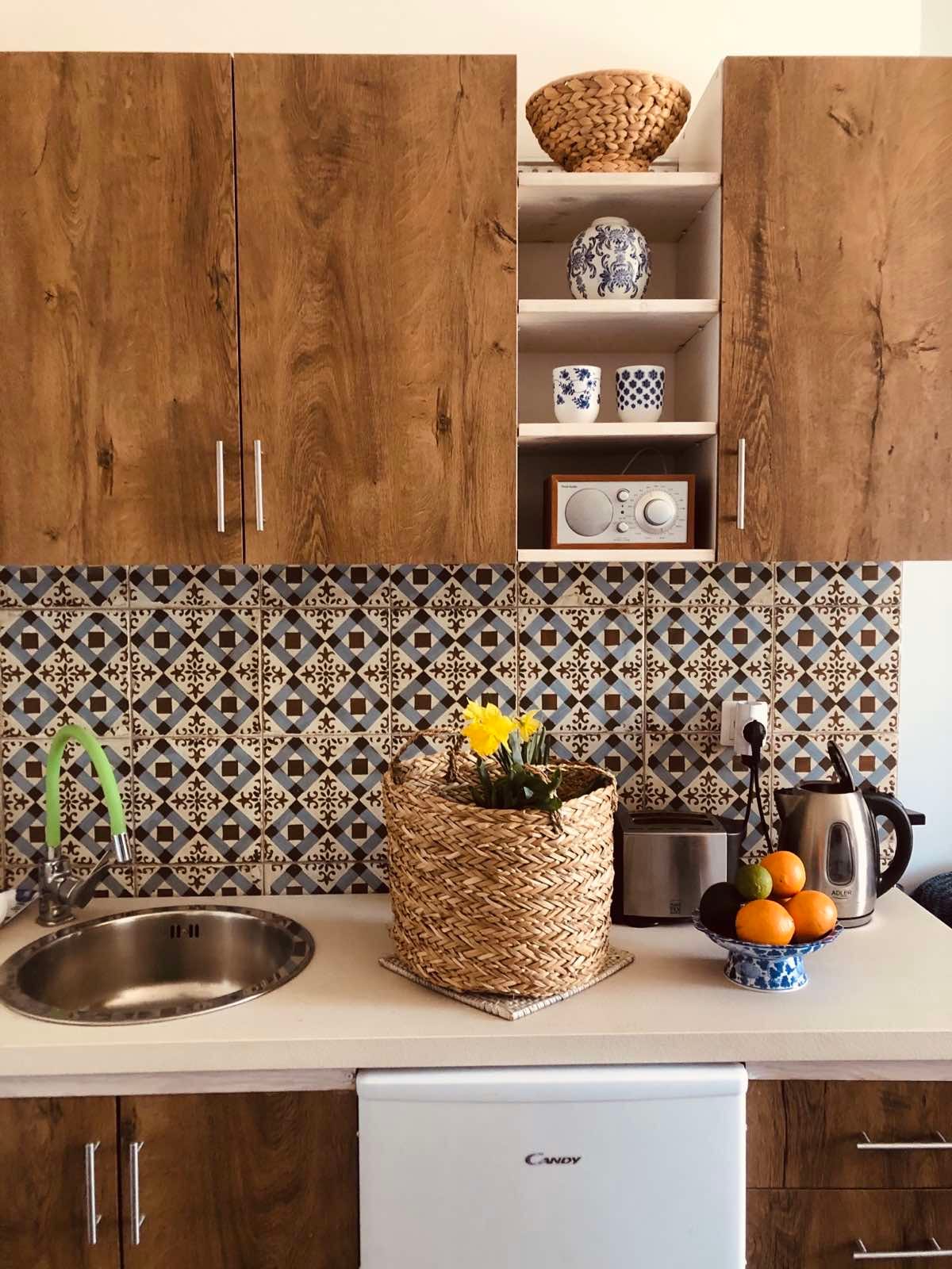 A compact kitchen area features wooden cabinetry and a decorative backsplash with geometric patterns. A stainless steel sink and appliances, including a kettle and a small refrigerator, are presented. A woven basket holds vibrant yellow flowers, while oranges rest in a blue patterned bowl.