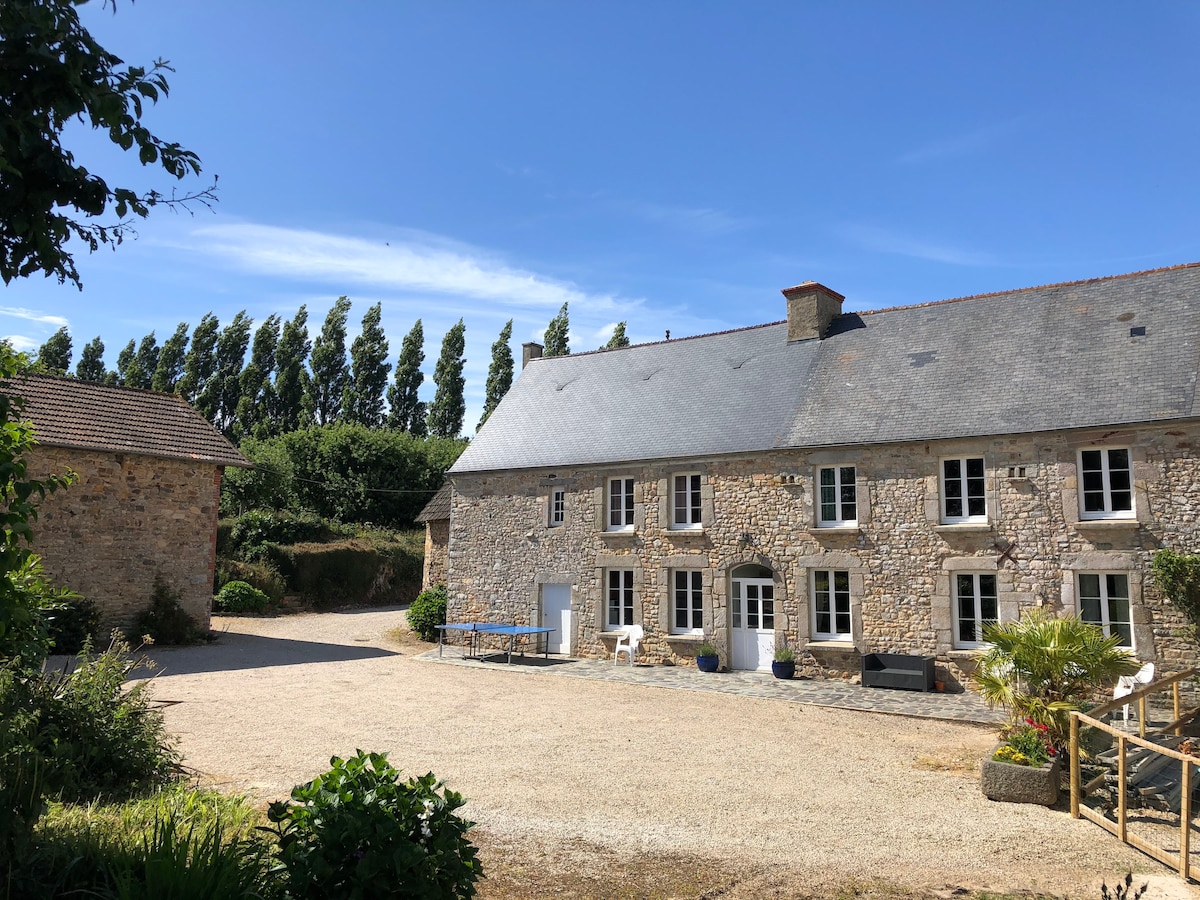 A charming stone farmhouse is showcased against a clear blue sky. The building features multiple windows and a slate roof, surrounded by lush greenery and well-maintained outdoor areas. A gravel path leads to the entrance, providing access to a serene courtyard.
