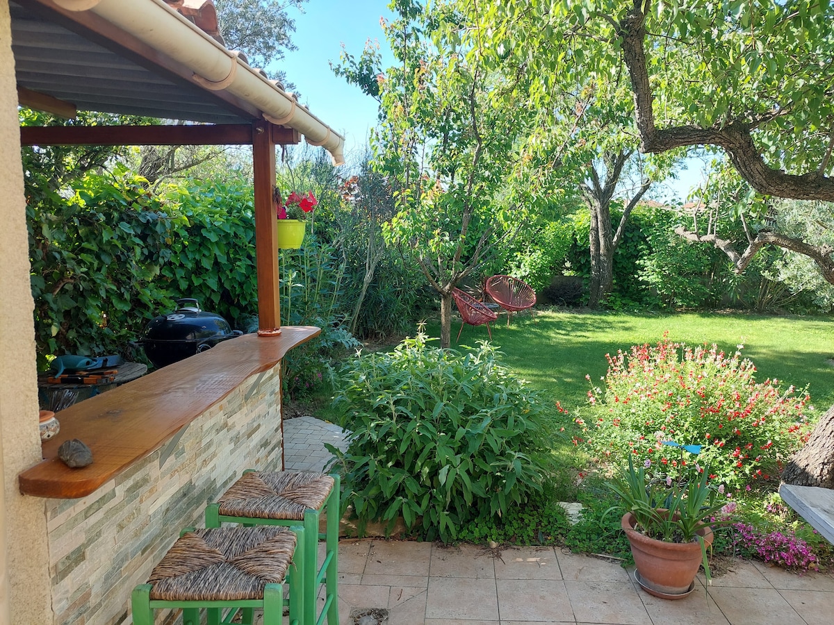An outdoor area features a wooden bar with stools, surrounded by lush greenery and colorful flowers. A grassy lawn is visible, along with a barbecue grill and a hammock, inviting relaxation in the tranquil garden atmosphere.