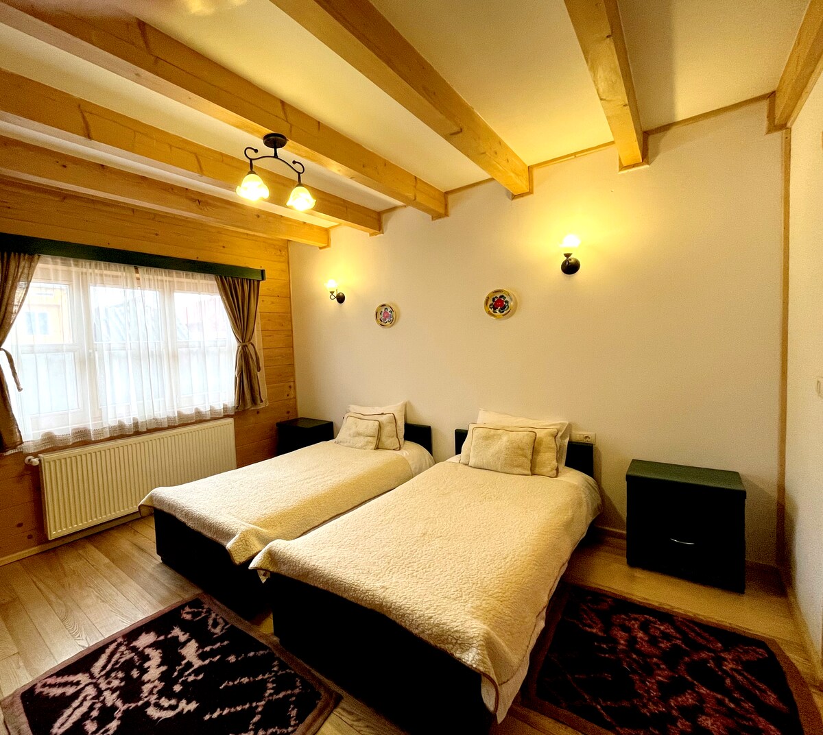A room is furnished with two spacious beds, each topped with soft white bedding. Natural wood beams line the ceiling, while a window dressed in light curtains allows soft light to enter. Two decorative plates adorn the wall, adding a touch of character.