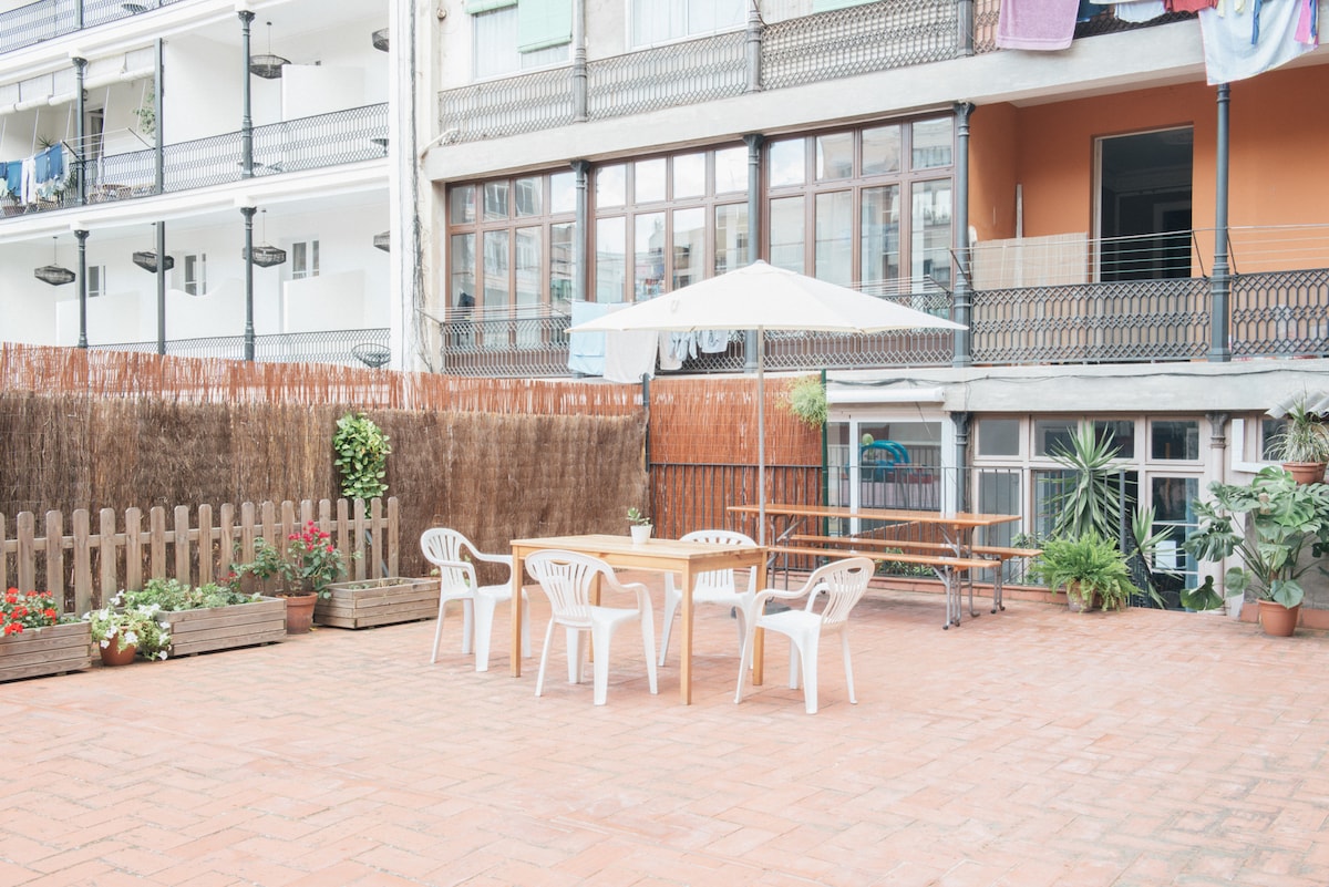 An outdoor patio area is shown, featuring a wooden table surrounded by four white plastic chairs. Potted plants and flowers are arranged along the edge, with a large umbrella providing shade. The backdrop includes several balconies adorned with laundry hanging to dry.