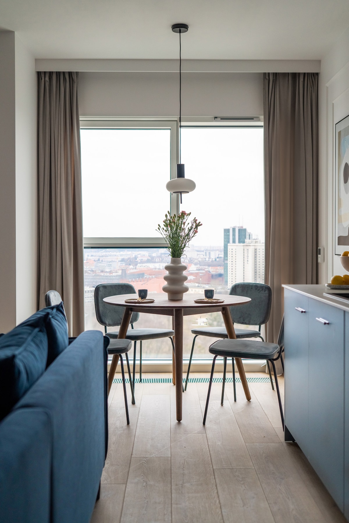 A modern dining area is shown with a round wooden table surrounded by two upholstered chairs. A decorative vase with flowers sits at the center. Large windows offer a view of the cityscape, while soft natural light filters through the light-colored curtains.