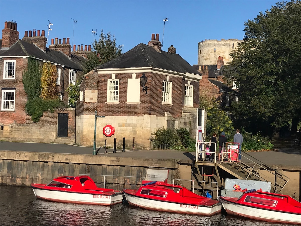 The historic boathouse, featuring red boats along the riverside, is nestled near the medieval Clifford's Tower. The building showcases a mix of brick and stone, while trees and greenery frame the scene, offering a glimpse of the vibrant river environment.