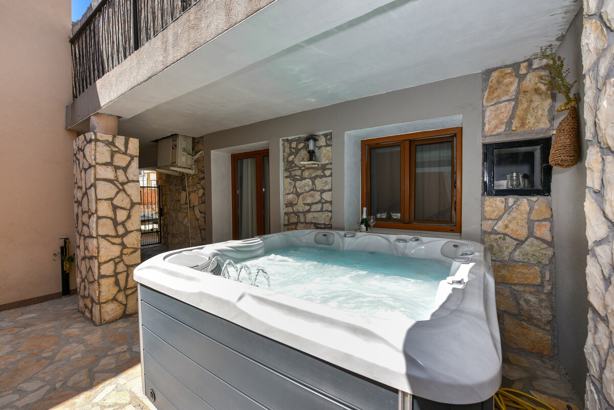 A private outdoor space features a jacuzzi designed for five people, surrounded by a stone facade. The area is partially shaded and includes windows offering views of the adjacent living space, enhancing the relaxing environment.