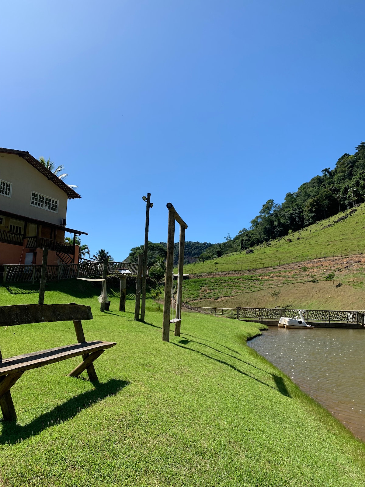 A scenic view of a well-maintained garden area with lush green grass sloping towards a calm body of water. A wooden bench is positioned on the grass, while a white pedal boat is visible on the water. The backdrop features a house with a balcony and surrounding trees.