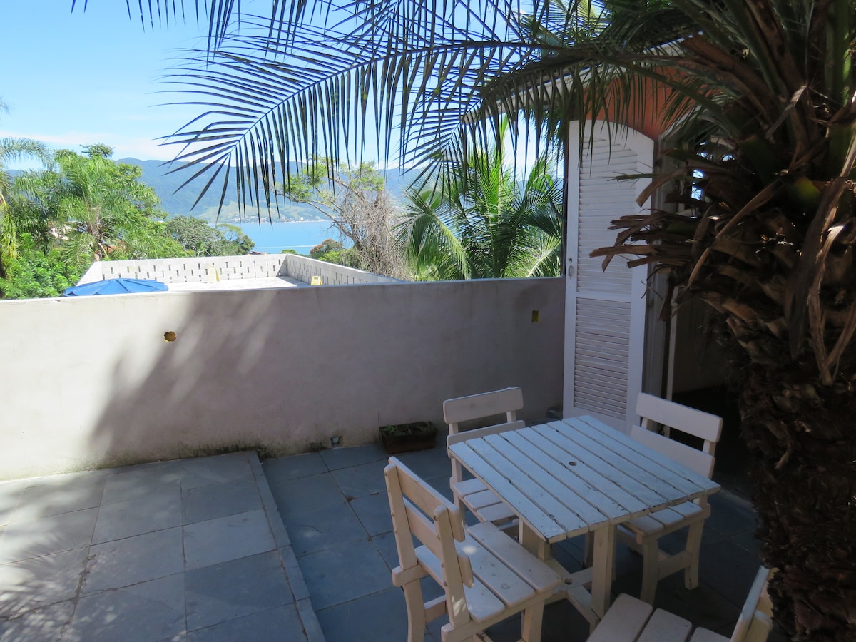 A small outdoor area is featured with a white wooden table and four chairs set on stone flooring. Lush palm trees surround the space, offering a natural setting while providing a view of the sea and nearby hills in the background.