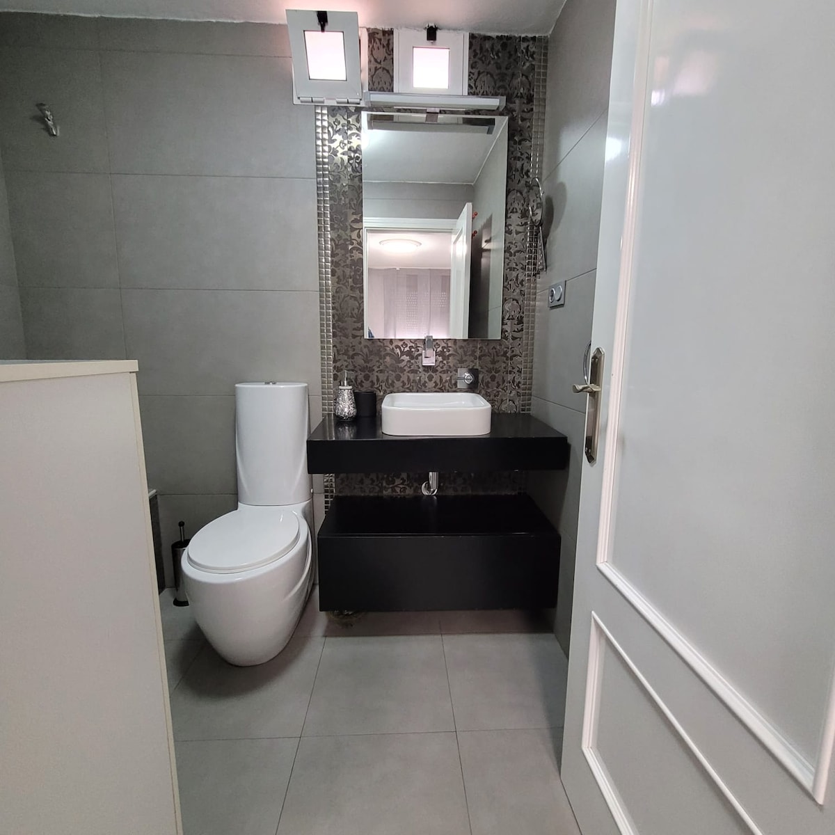 A modern bathroom features sleek grey tile flooring and wall tiles. A white toilet and a minimalistic sink are complemented by a dark countertop. Clean lines and stylish lighting add to the contemporary design, creating a functional and well-appointed space.