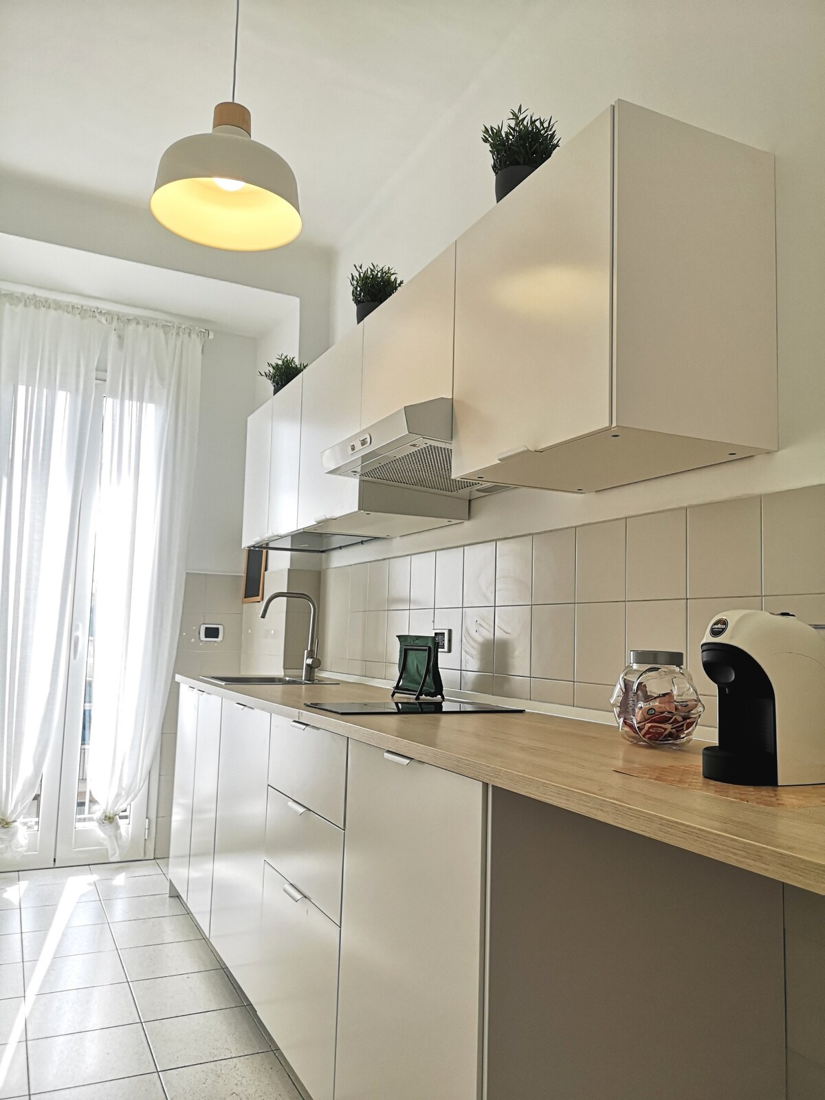 A modern kitchen features sleek cabinetry and a wooden countertop. Bright light filters through the sheer curtains, enhancing the bright ambiance. A coffee machine and a clear jar of snacks are positioned on the counter, with small plants adorning the upper shelves.