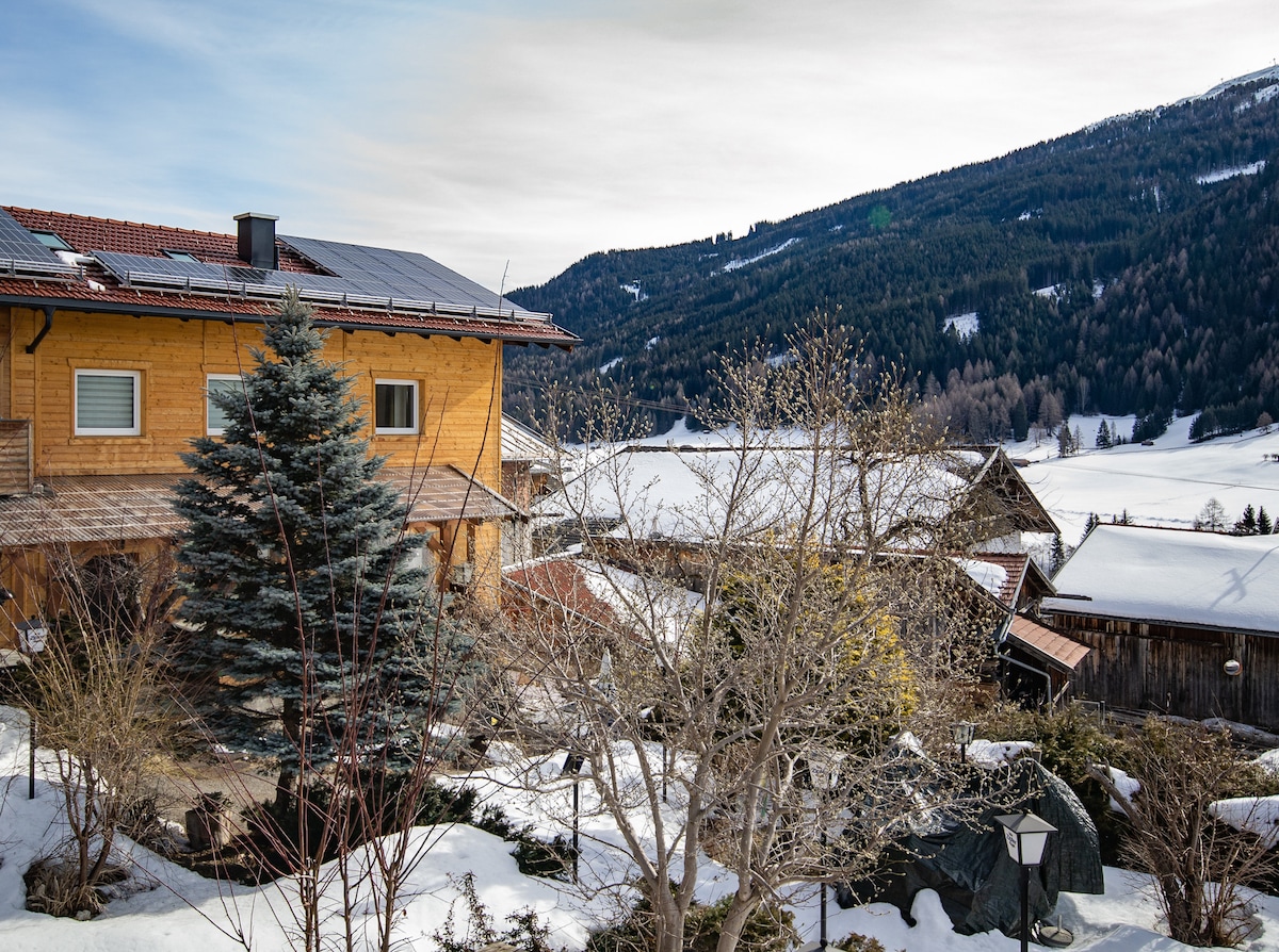 A scenic view of a wooden house surrounded by snow-covered landscapes. Solar panels are visible on the roof, and several trees dot the foreground. The mountains rise in the background, partially shaded by clouds, creating a tranquil winter atmosphere.