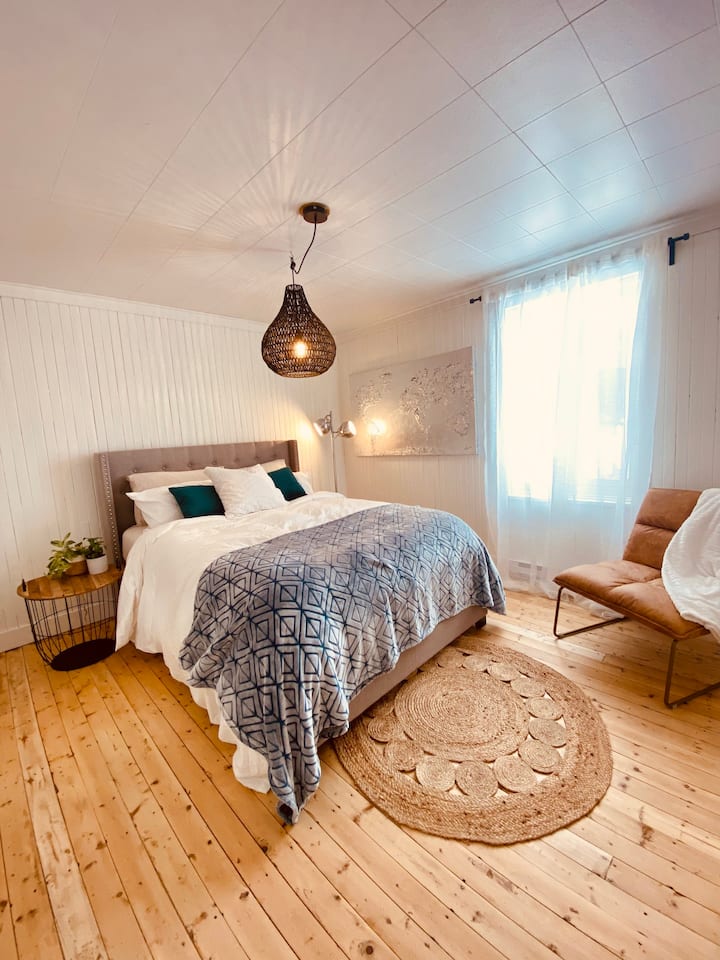 Un lit Queen douillet dans une chambre baignée de lumière, avec fauteuil d’appoint et commode pour un séjour confortable.