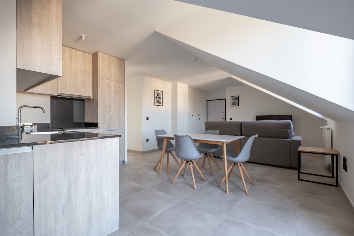 An open-concept living area is revealed, featuring a modern kitchen with wooden cabinetry, next to a dining table surrounded by gray chairs. A spacious sofa is positioned against the wall, with natural light filtering through a triangular window above.