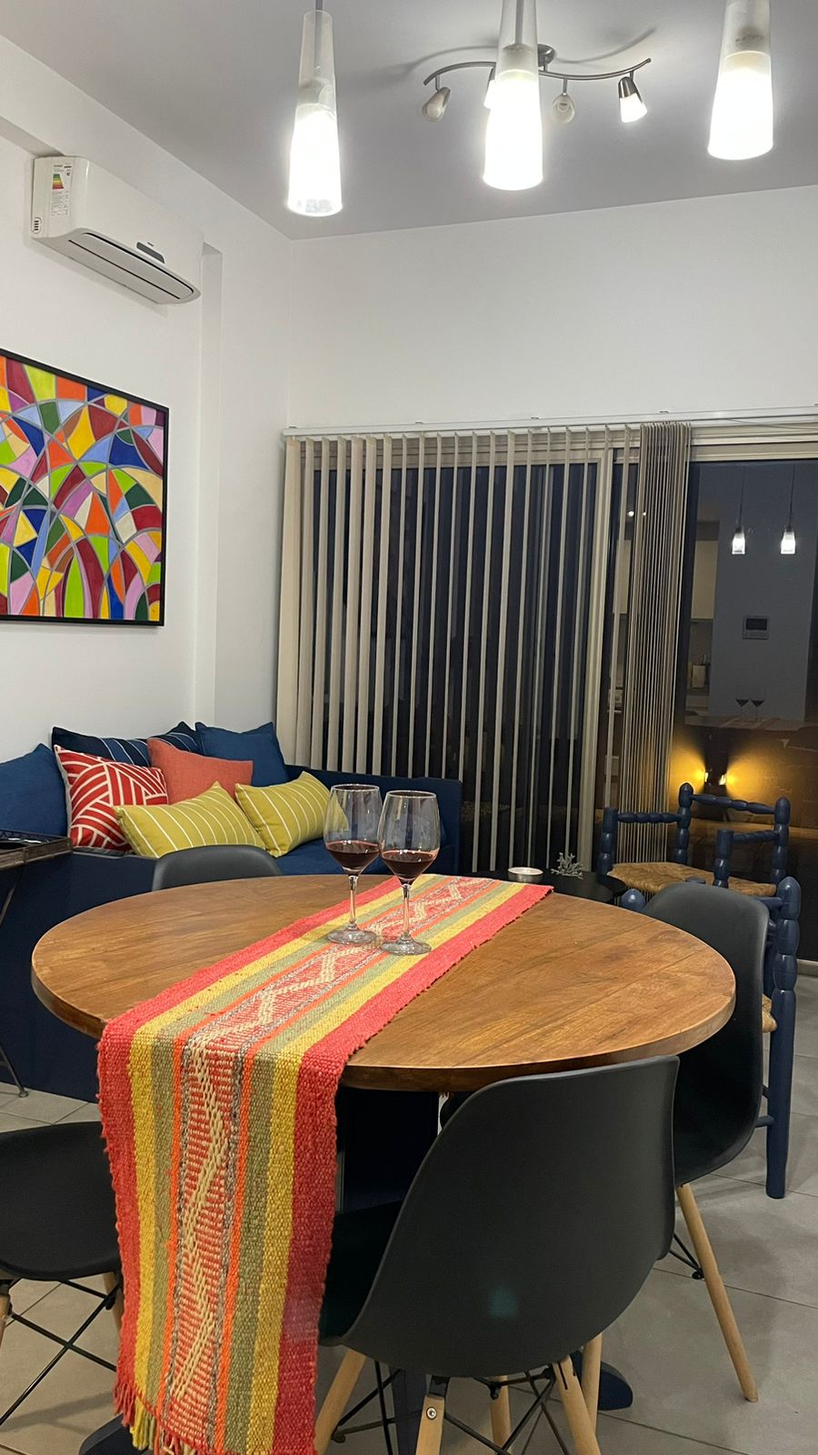A dining area is presented with a round wooden table surrounded by black chairs. Colorful cushions are arranged on a nearby seating area. A vibrant table runner adds a touch of color, and large windows with vertical blinds allow natural light to fill the space.