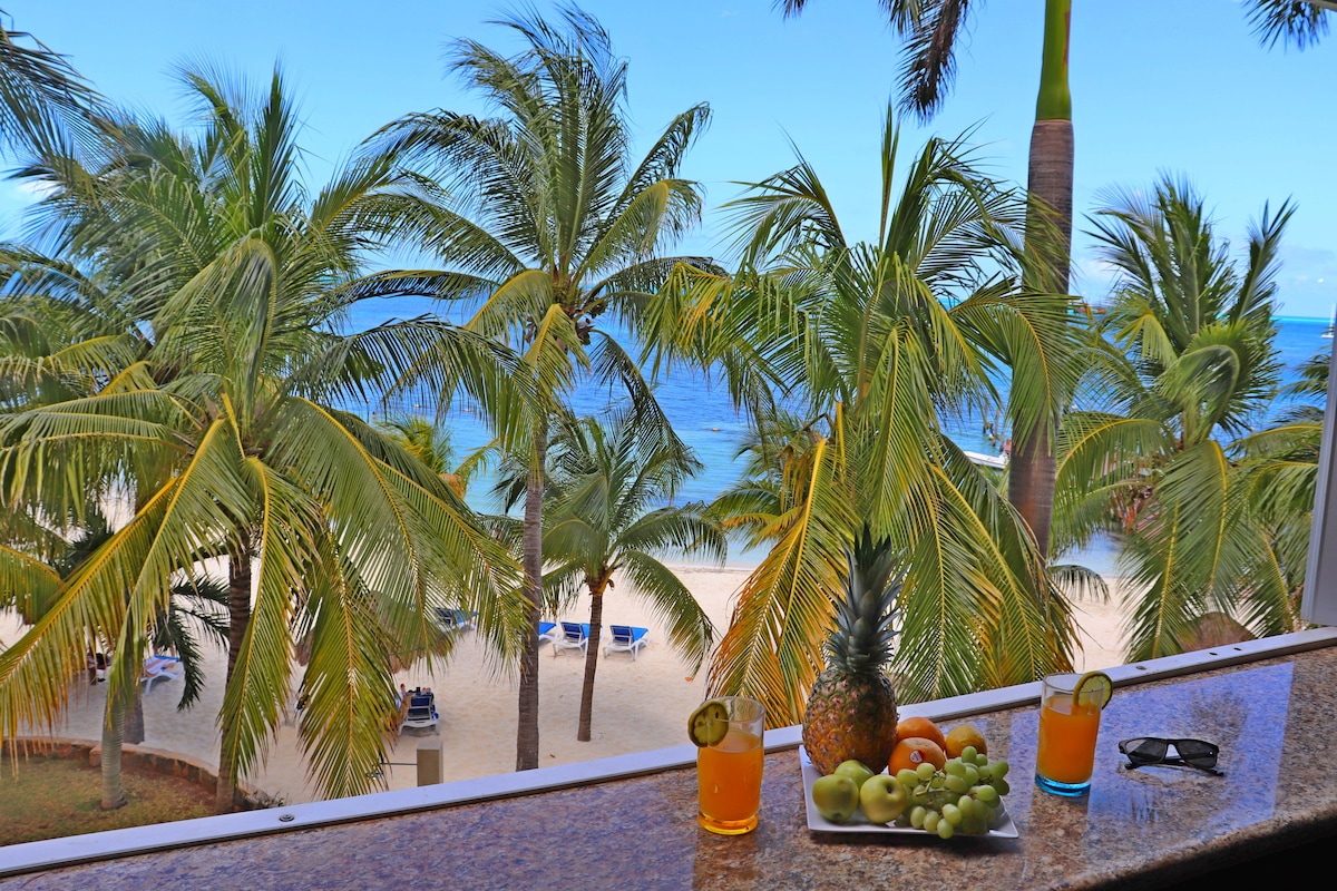 A balcony view displays vibrant palm trees swaying in the breeze, framing a serene beach and ocean beyond. A tray of fresh fruits and tropical drinks sits on the ledge, inviting relaxation while enjoying the coastal scenery.