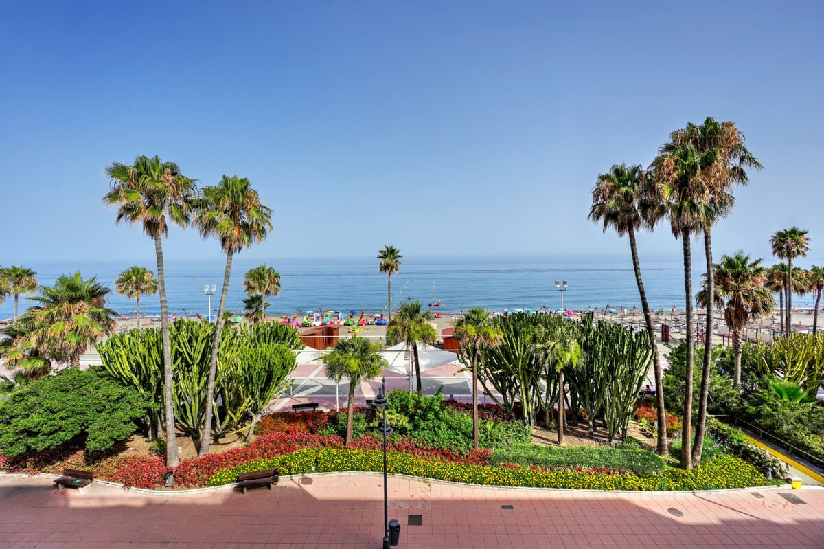 The image displays lush greenery and vibrant flowers lining a coastal pathway. Palm trees sway gently in the breeze, while the calm sea extends to the horizon. Sunlight brightens the scene, casting a serene atmosphere during daytime, with beachgoers visible in the distance.