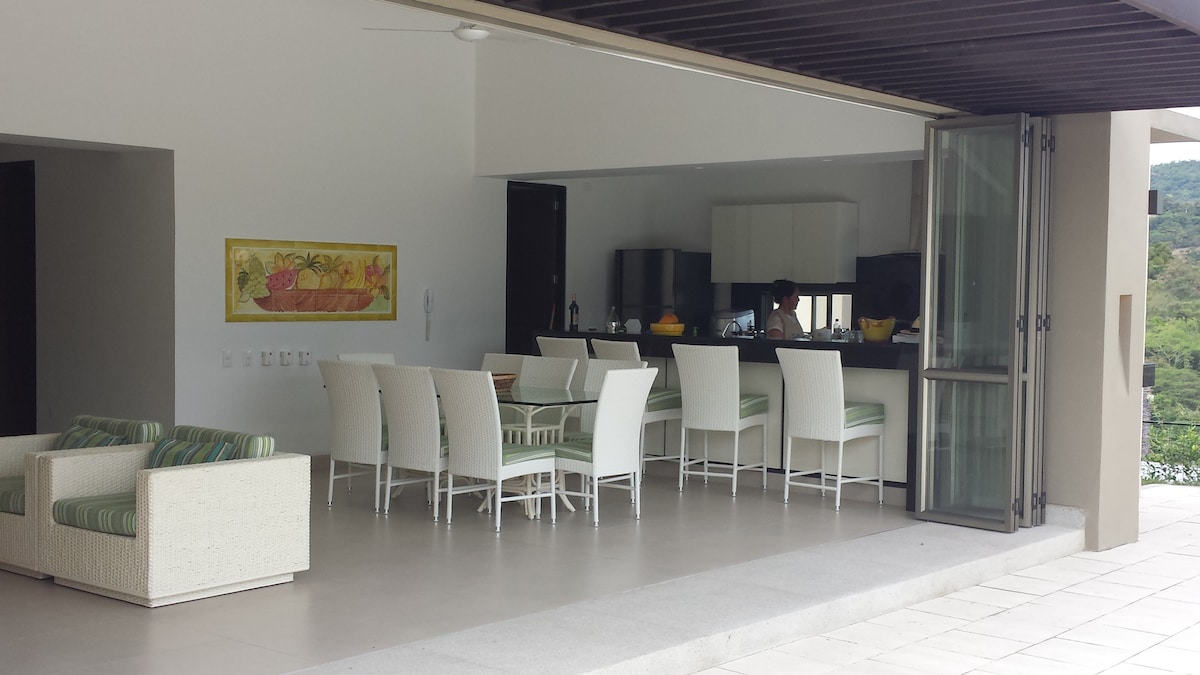 An open-concept dining area features a large table surrounded by white chairs. A view of the kitchen is visible, where meal preparations can be observed. Natural light enters through expansive glass doors that lead to an outdoor area, with a subtle greenery view in the background.