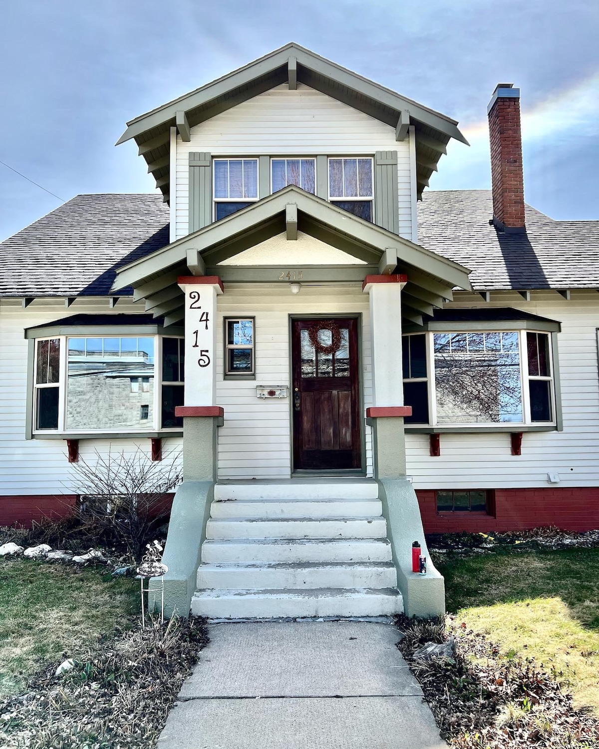 A historic craftsman-style home is presented, featuring a welcoming porch with steps leading to a central front door. Decorative trim accentuates the roofline, and large windows allow ample natural light. A well-maintained lawn and garden border the pathway, enhancing the home's curb appeal.