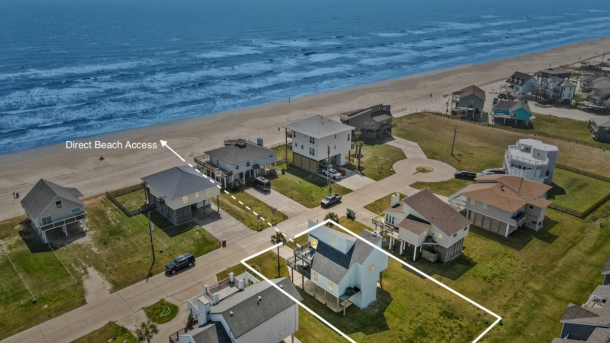 An aerial view highlights the coastal neighborhood with direct beach access. The surrounding homes are positioned along a sandy shoreline, with the ocean waves gently breaking nearby. Green grass and open spaces are visible, inviting outdoor exploration.