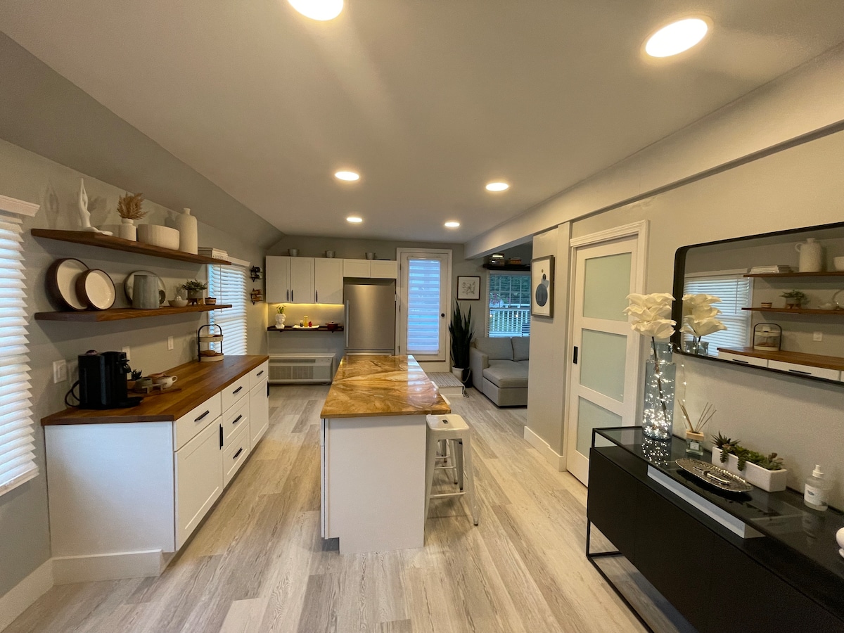 A modern kitchen is observed, featuring white cabinetry and a wood-topped island with seating. Open shelving holds decorative dishware. Stainless steel appliances complement the space, while ample lighting enhances the functionality. A sleek black console table displays ornamental items and greenery.