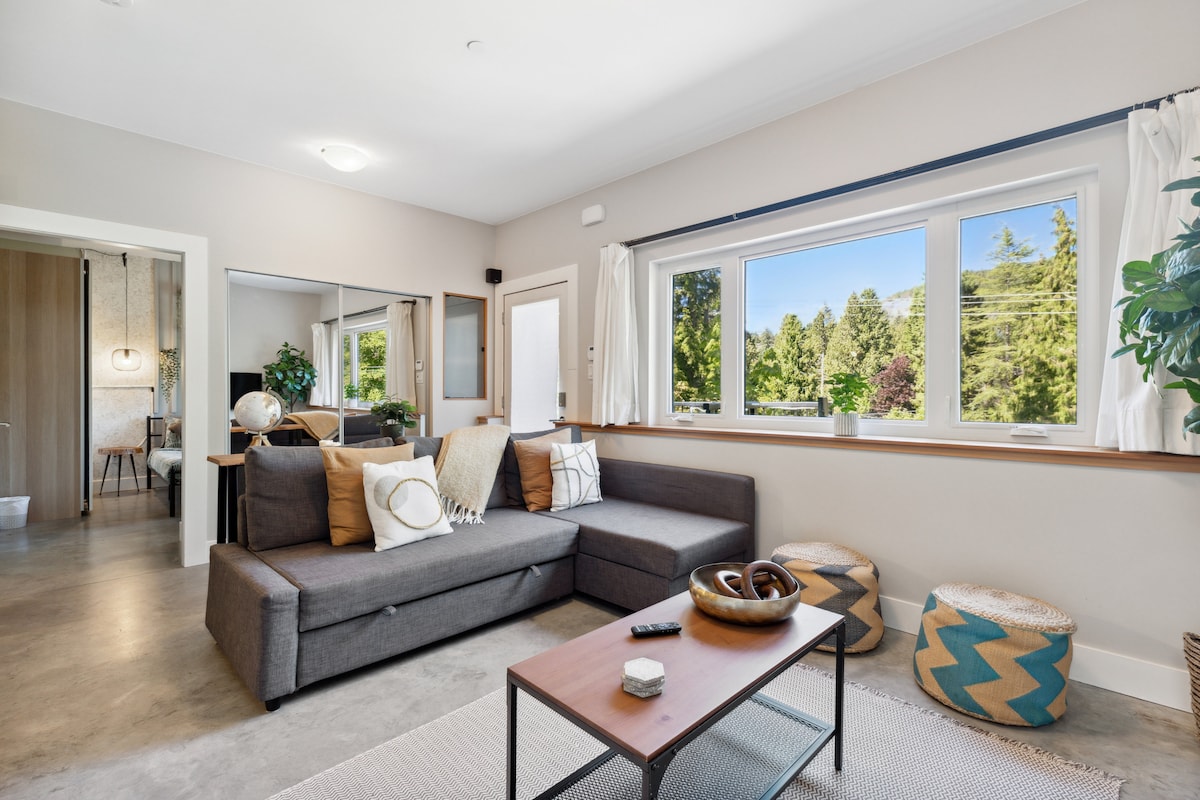 The living area features a spacious gray sofa positioned against a large window, offering views of the surrounding greenery. A wooden coffee table sits in the center, complemented by decorative items. Soft natural light illuminates the room, creating a calm environment.