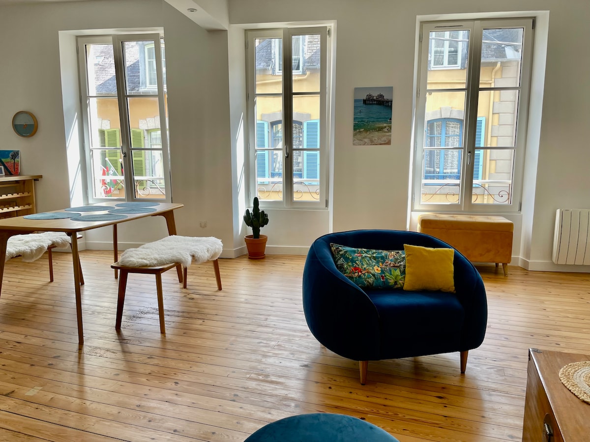A bright living area features large windows allowing natural light to fill the space. A round, blue armchair is positioned beside a wooden table, which is accompanied by cozy sheepskin covers. A small potted plant adds a touch of greenery to the wooden floor.
