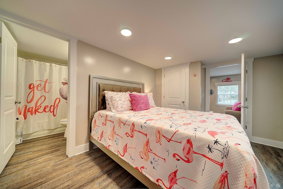 A cozy bedroom featuring a spacious bed adorned with a colorful flamingo-patterned quilt. A subtle arrangement of bright pink pillows adds a playful touch. The bathroom is accessible through a door, and natural light enters from a nearby window, enhancing the inviting atmosphere.