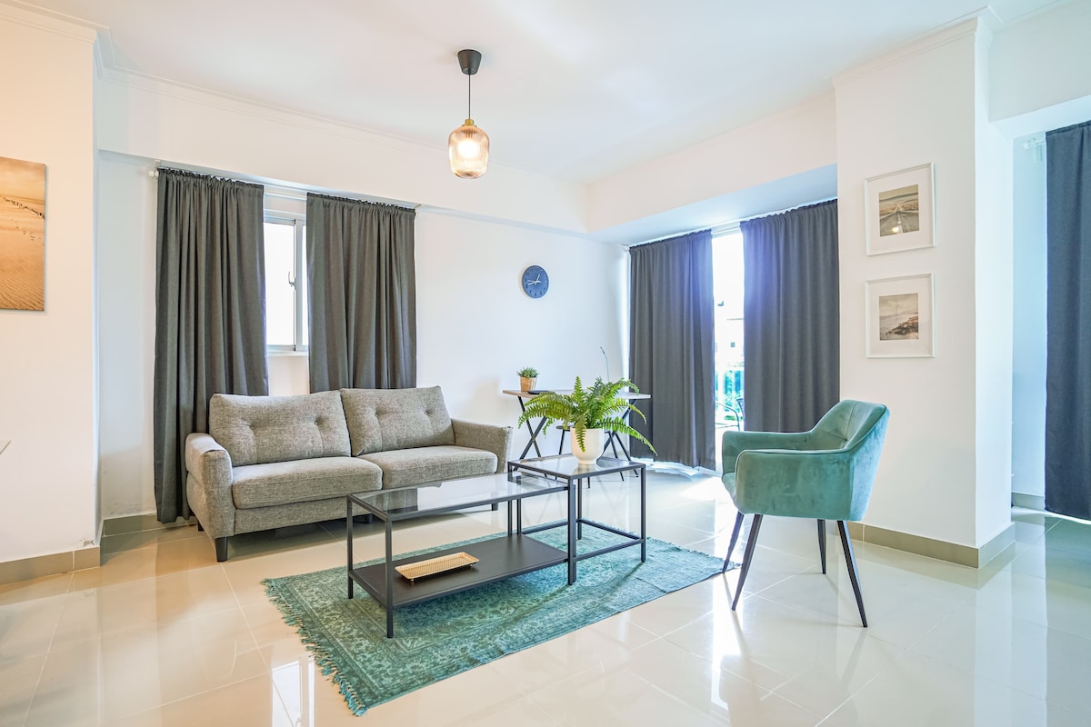 The living room features a grey sofa and a teal armchair, arranged around a low black coffee table on a patterned rug. Natural light enters through large windows, complemented by dark curtains. A small dining table is visible in the background, alongside decorative wall art.