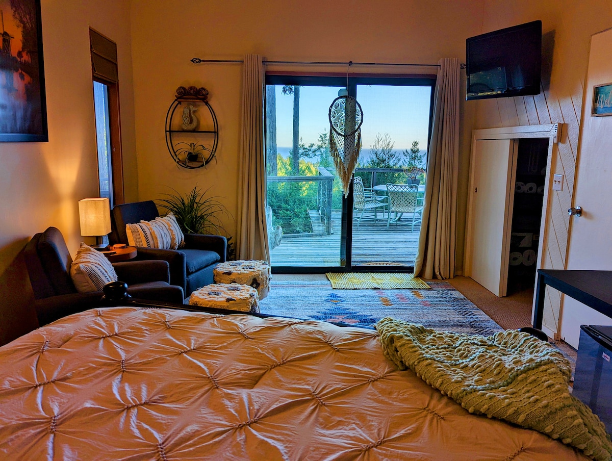 A cozy sleeping area features a spacious bed with an inviting duvet and textured throw. A seating area with two chairs is visible next to a small table. Large glass doors open to reveal a private deck with outdoor seating and an expansive view.