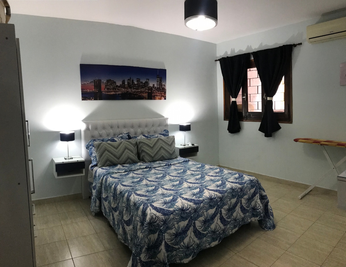 A well-lit bedroom features a spacious bed dressed in a blue and white patterned coverlet. Nightstands with lamps flank the bed, providing additional lighting. A large cityscape canvas hangs above the headboard. Black curtains frame the window, while an ironing board is positioned nearby.