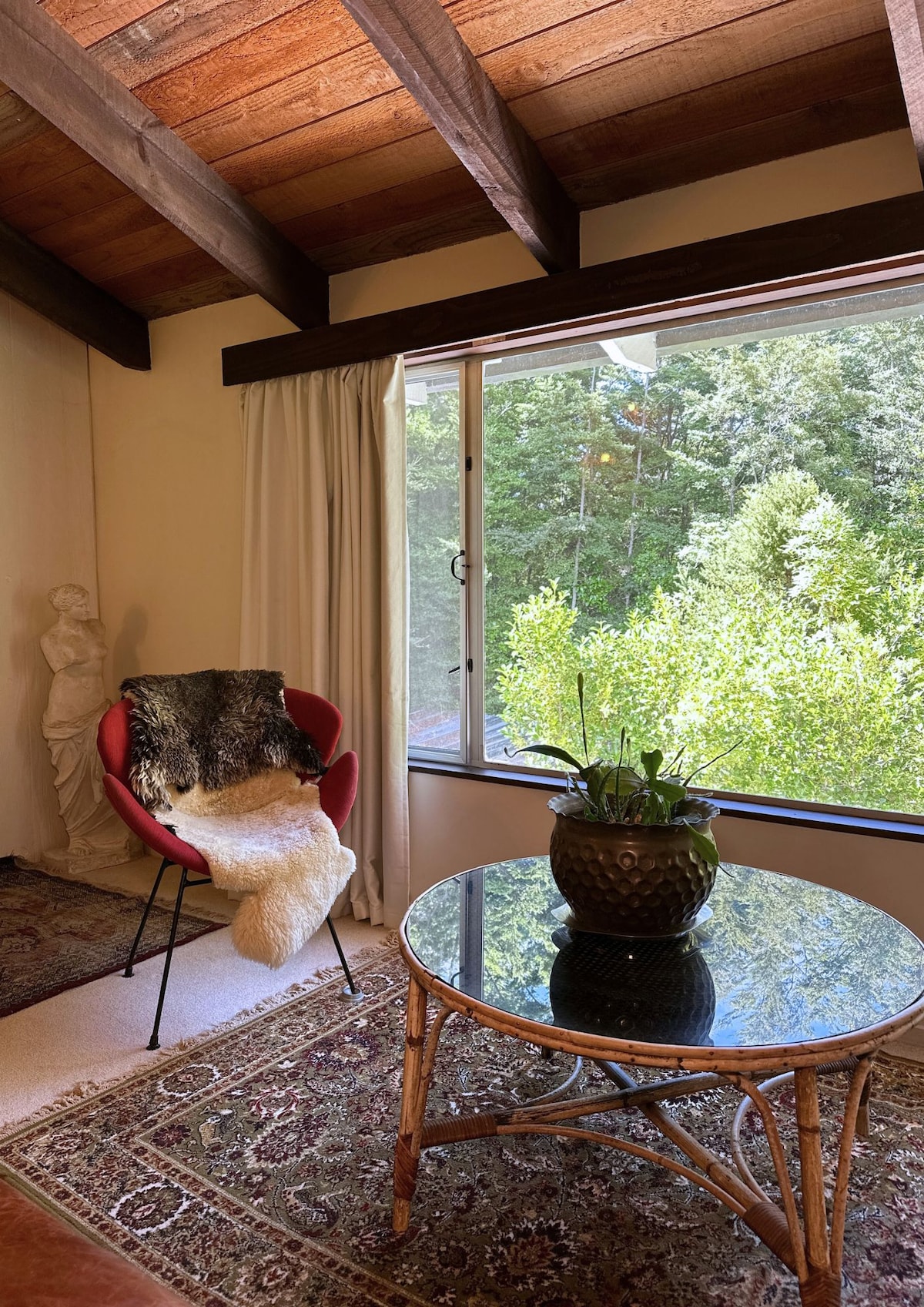 A cozy corner is highlighted by a circular glass table resting on a patterned rug. A red chair, adorned with a soft throw, is positioned near a large window that offers views of the surrounding lush greenery, enhancing the serene ambiance.