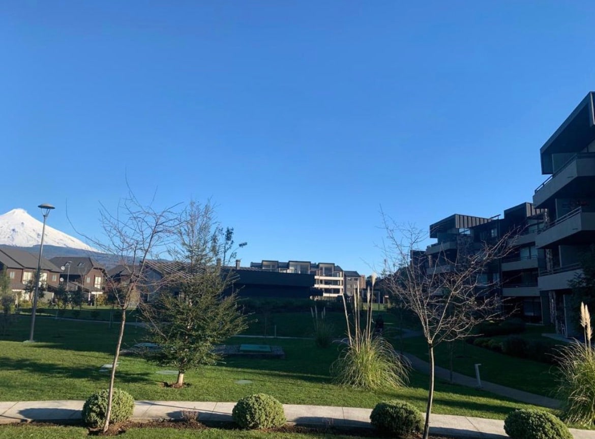 A clear blue sky frames the scene, depicting a landscaped area featuring manicured grass and small trees. Several modern buildings are visible, complemented by distant mountain views. Pathways and green spaces create an inviting outdoor environment.