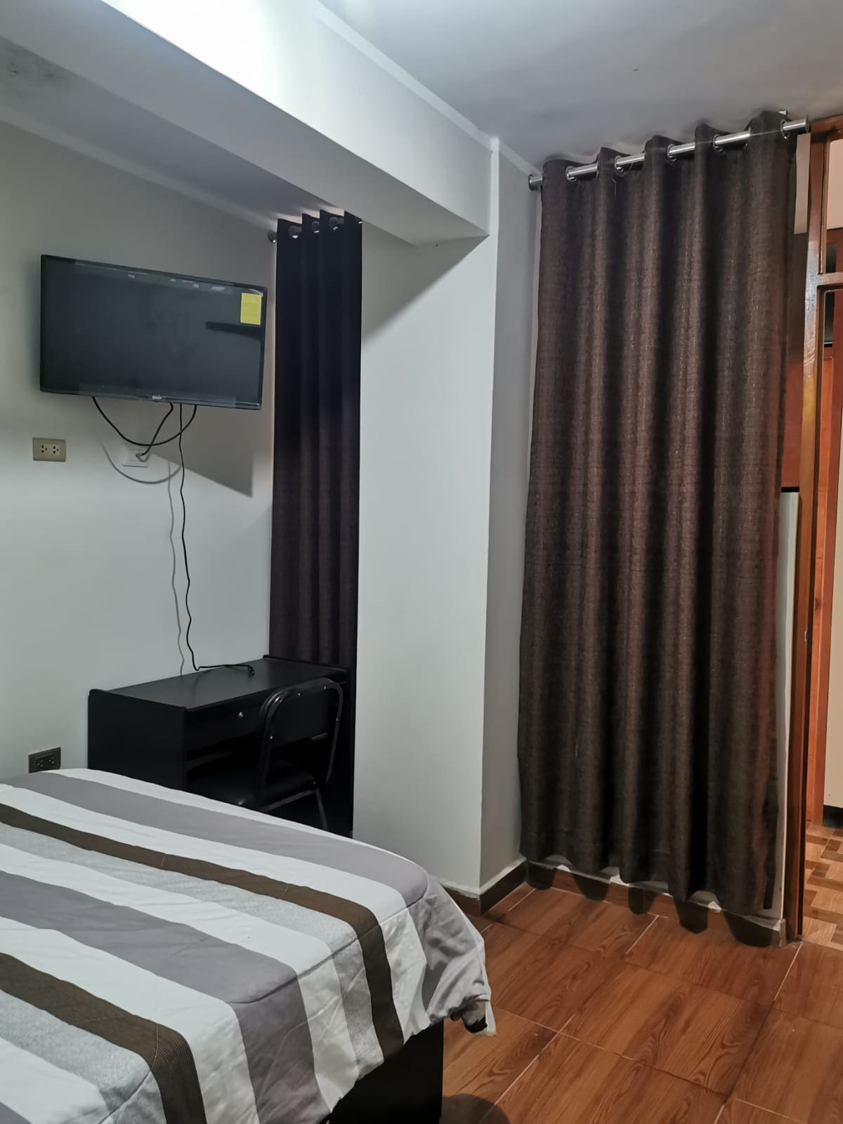 A cozy bedroom features a spacious bed covered with a striped blanket. A wall-mounted television is positioned above a desk accompanied by a chair. Dark curtains frame the window, and a closet can be seen in the background, providing additional storage.