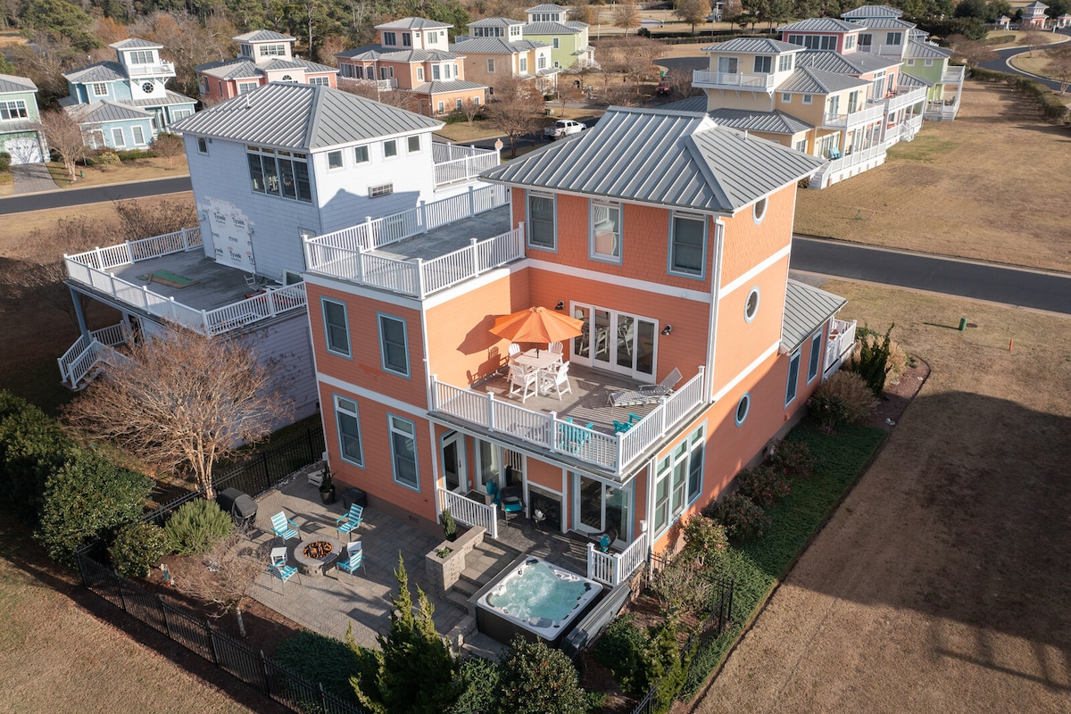 The exterior of a vibrant orange house is shown, featuring multiple balconies and a spacious patio area with outdoor seating. A hot tub is visible on the patio, surrounded by landscaping. Nearby, additional homes can be seen, highlighting the neighborhood's character and layout.